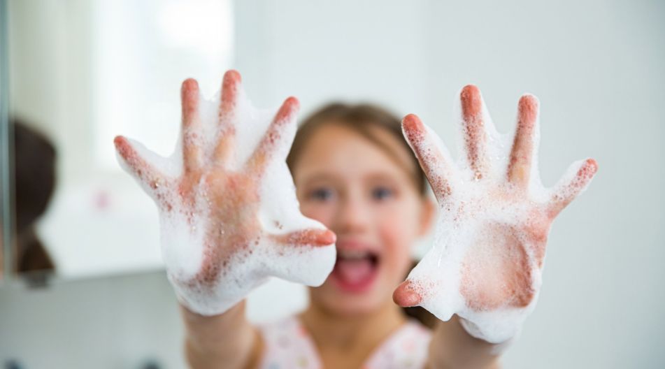 Ein Kind hält lachend seine mit Schaum bedeckten Hände in die Kamera, während es sich die Hände wäscht.