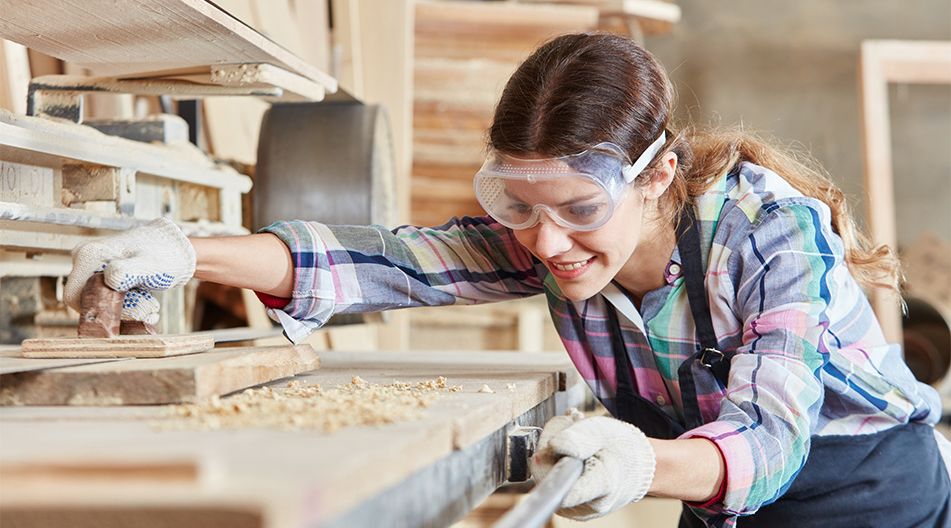 Frau mit Schutzbrille und Handschuhen bearbeitet konzentriert ein Holzstück in einer Werkstatt an einer Schleifmaschine.
