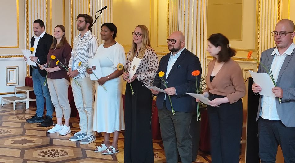 Gruppe von Erwachsenen steht in einem festlichen Raum in einer Reihe und hält Urkunden sowie Blumen in den Händen.