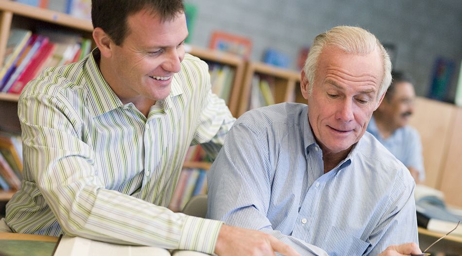 Ein jüngerer Mann hilft einem älteren Mann beim Lernen in einer Bibliothek. Beide tragen Hemden und schauen gemeinsam in ein aufgeschlagenes Buch, während sie lächeln.