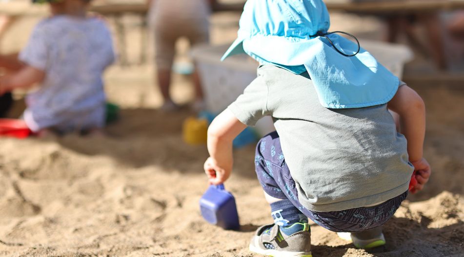 Kleinkind mit Sonnenhut spielt im Sandkasten und gräbt mit einer Schaufel, während andere Kinder im Hintergrund sind.