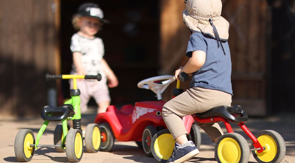 Ein Kind mit Sonnenhut sitzt auf einem roten Dreirad, während ein weiteres Kind mit Baseballkappe im Hintergrund steht. Mehrere bunte Kinderfahrzeuge stehen auf einem sonnigen Hof.