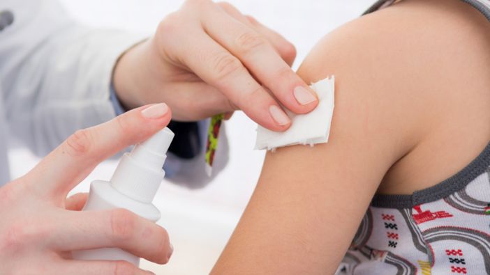 Eine Person desinfiziert den Oberarm eines Kindes mit einem weißen Tuch und einer Sprühflasche, vermutlich vor einer medizinischen Behandlung oder Impfung. Eine Person desinfiziert den Oberarm eines Kindes mit einem weißen Tuch und einer Sprühflasche, vermutlich vor einer medizinischen Behandlung oder Impfung.