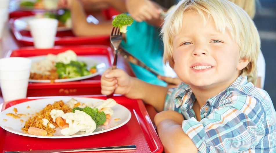 Ein Grundschüler sitzt am Mittagstisch, grinst mit einem auf einer Gabel aufgespießten Brokkoli in die Kamera. Auf dem Teller vor ihm liegt weiteres Gemüse und Reis.