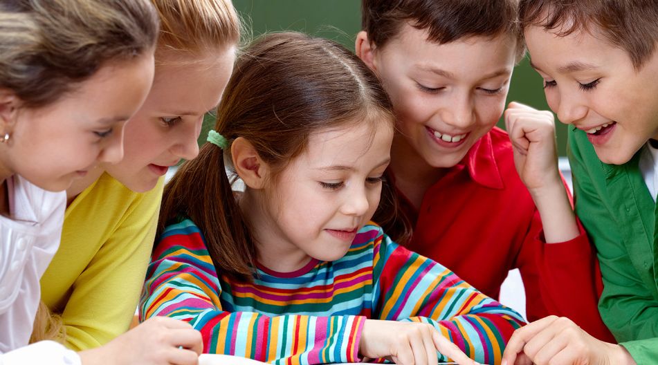 Eine Gruppe von fünf fröhlichen Grundschulkindern sitzt zusammen und liest in einem aufgeschlagenen Buch. Sie schauen gespannt auf die Seiten.