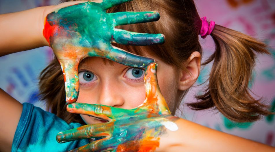 Ein Kind mit blauen Augen und braunen Zöpfen hält bemalte Hände vors Gesicht und formt mit den Fingern einen Rahmen, durch den es schaut. Die Hände sind bunt mit Farben bedeckt, im Hintergrund sind Handabdrücke an der Wand zu sehen.